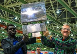 A student and a teacher holding a trophy together inside of a factory.