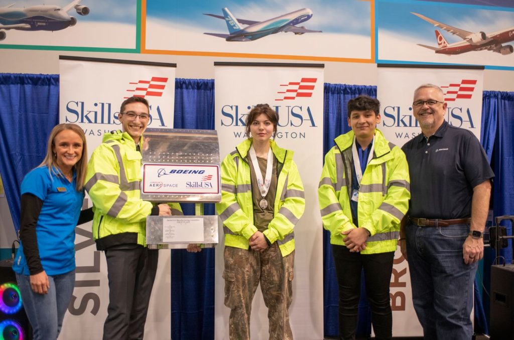 A photo of three students and one student is holding a trophy. Two adults ore on both sides of the student group. 