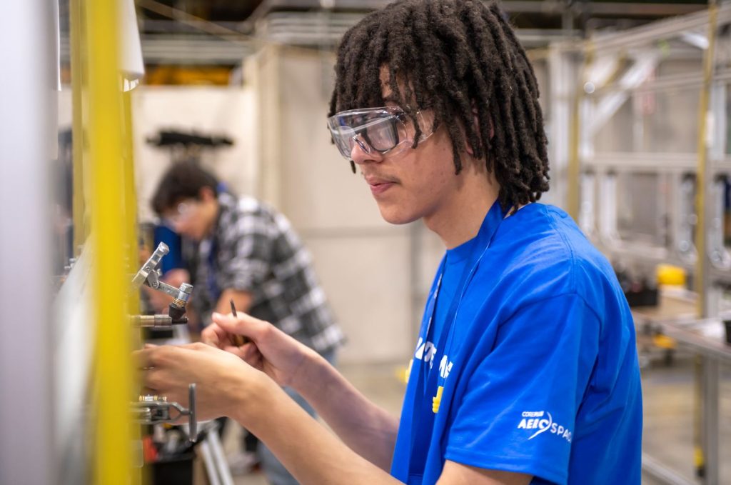 Photo of a student working on a hands-on project inside of factory. The student is wearing safety glasses and a blue shirt.