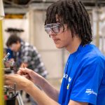 Photo of a student working on a hands-on project inside of factory. The student is wearing safety glasses and a blue shirt.