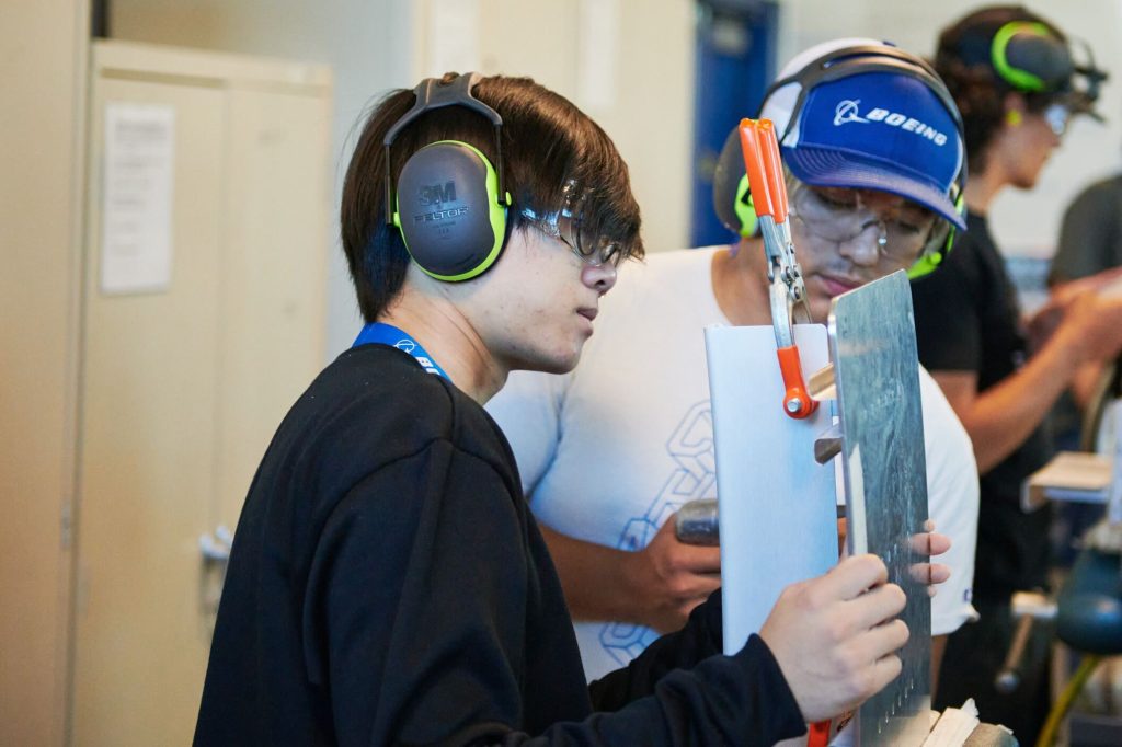 Two students working on a hands-on project with a piece of metal. Both students are inside of training facility and wearing hearing and eye protection.