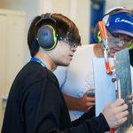 Two students working on a hands-on project with a piece of metal. Both students are inside of training facility and wearing hearing and eye protection.