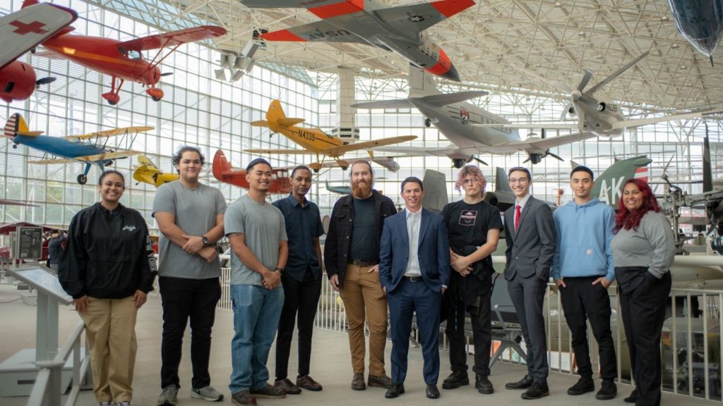  A group photo of people standing in a museum. Behind the group are planes. 