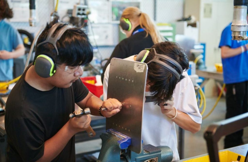 Two students working on a hands-on project using hand tools. They are working on a piece of metal in a vice. They are in a training facility.